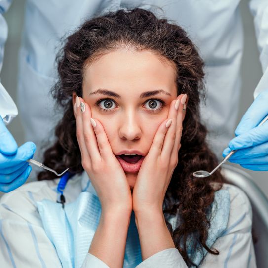 A woman in a dental chair showing a concerned expression with hands on her face.