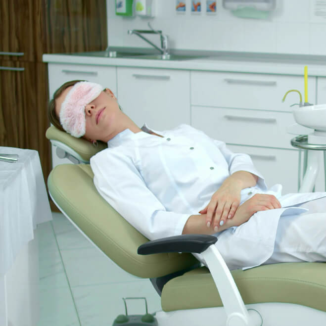 A woman wearing an eye mask reclining in a dental chair in a clinical setting.